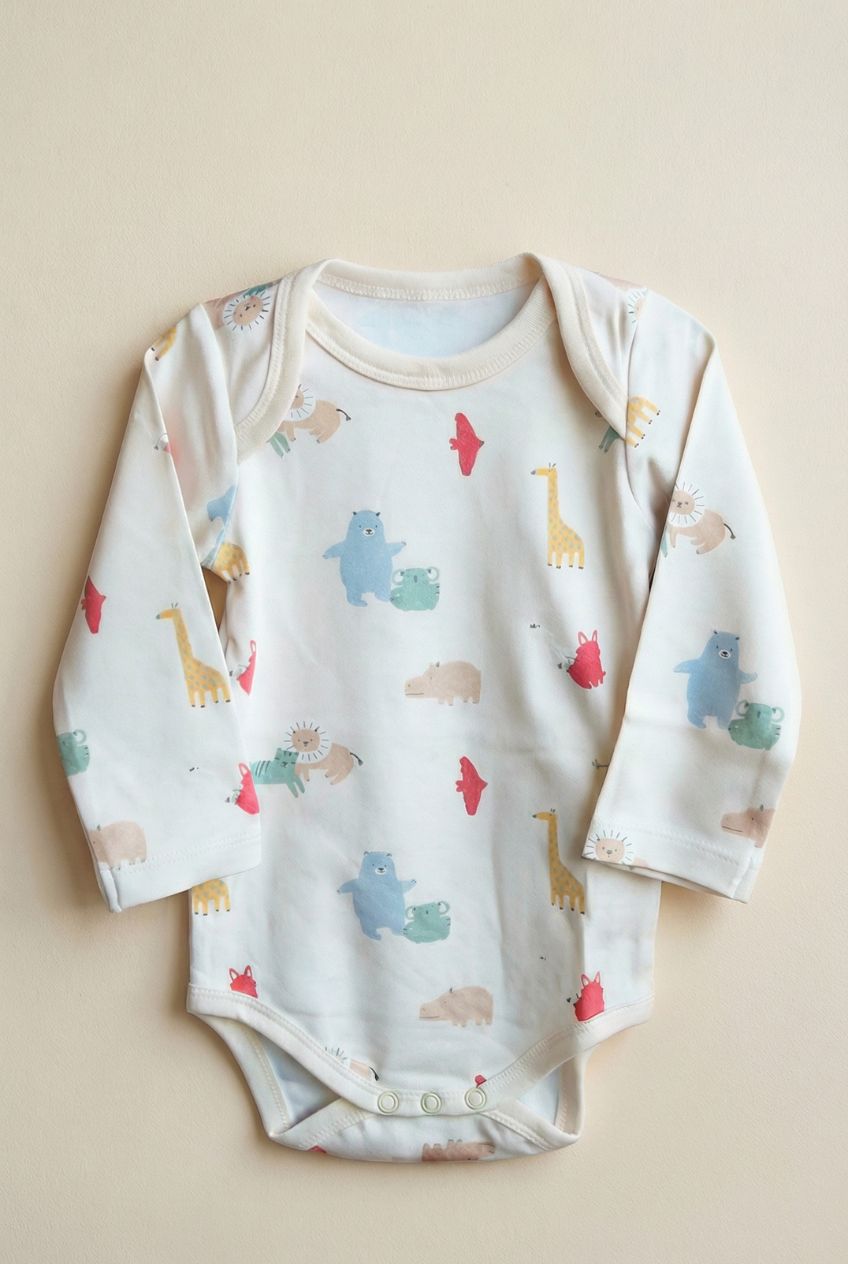 Ekologisk babybody från kollektionen Safari Vänner med färgglatt djurmönster på vit botten, flatlay på beige bakgrund